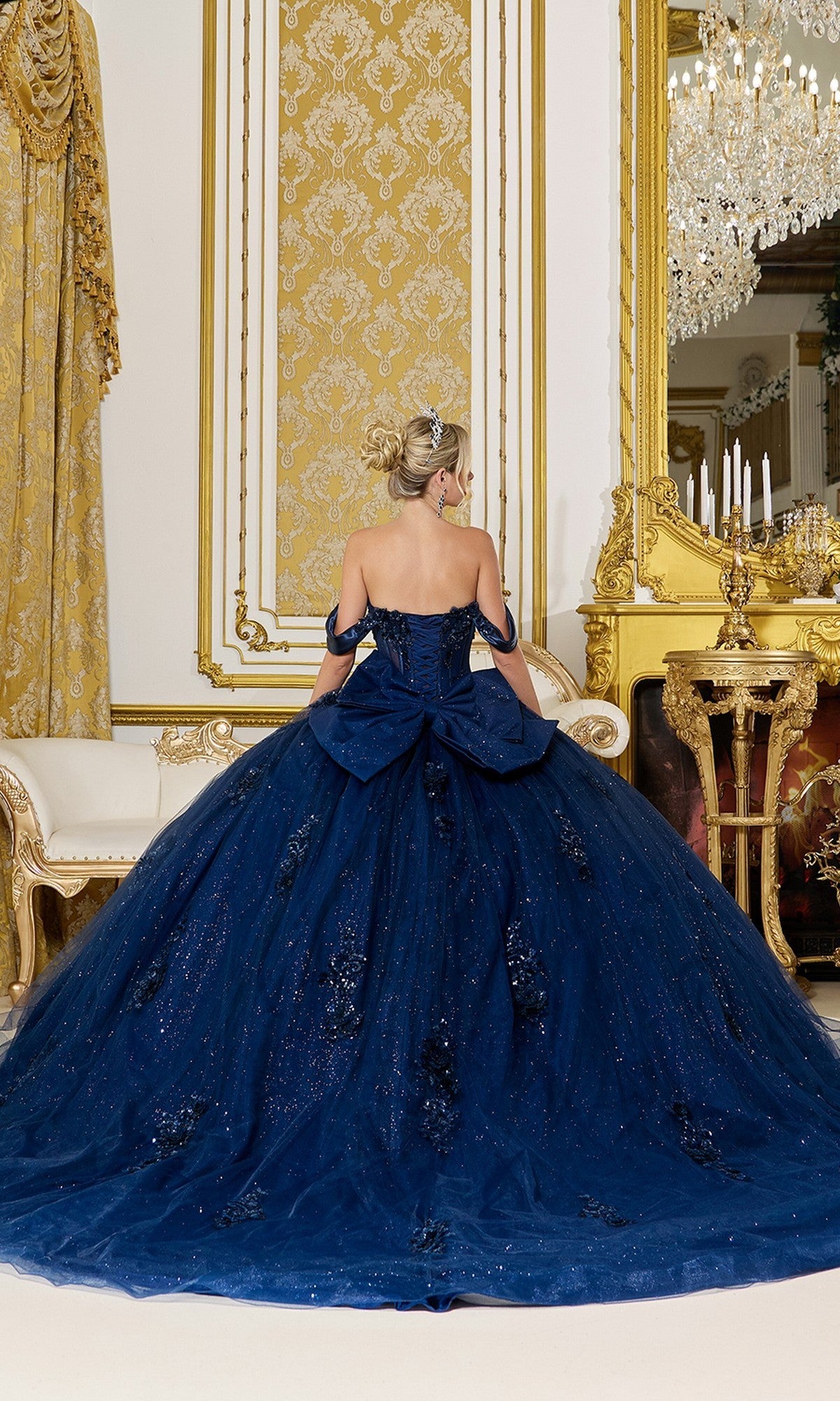 A woman wearing the Quinceanera Dress 5101 By Dancing Queen, featuring navy blue fabric, beaded applique, and a large bow, stands facing away in an ornate gold-accented room with a chandelier and classic furniture.